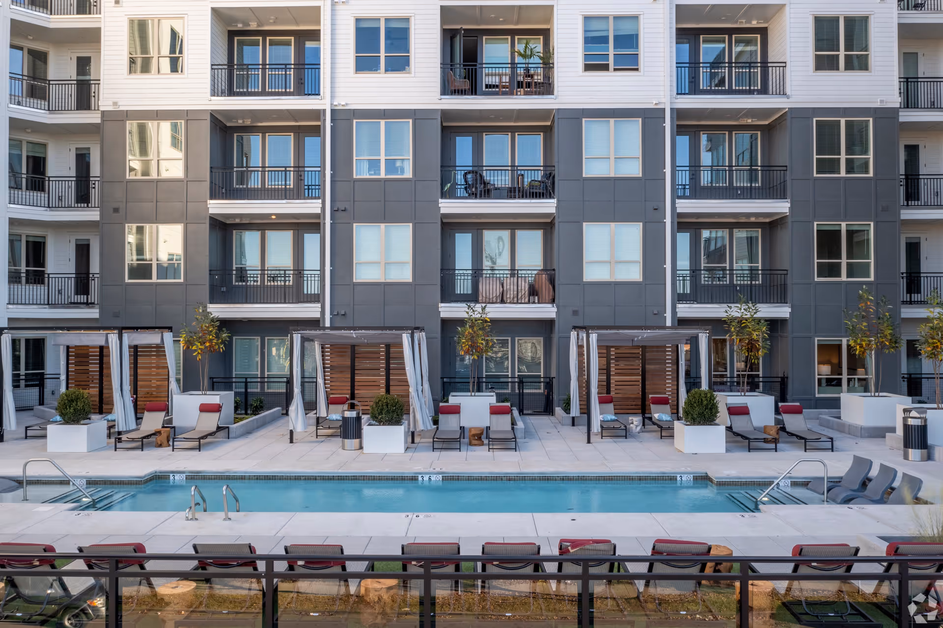 Modern multi-story apartment building with balconies overlooking a courtyard swimming pool and lounge cabanas.