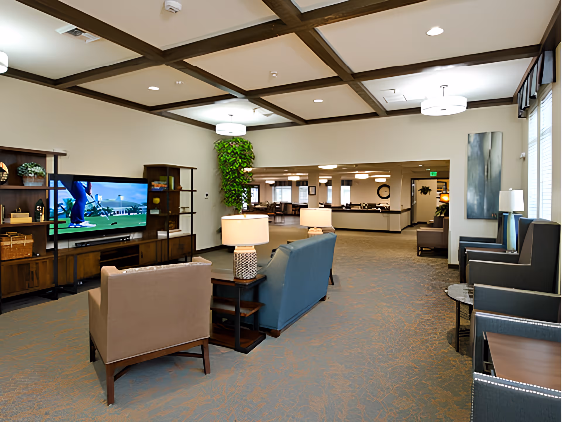 Spacious senior living common area with seating, a large TV, lamps, and a reception area visible in the background.