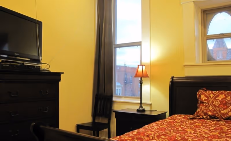A bedroom with a dark wood bed and dresser, red patterned bedding, a TV on the dresser, a bedside lamp, and two windows.
