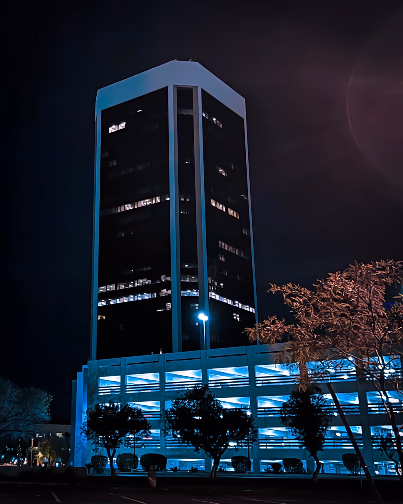 Nighttime exterior view of a tall, blue-lit building with a parking structure.