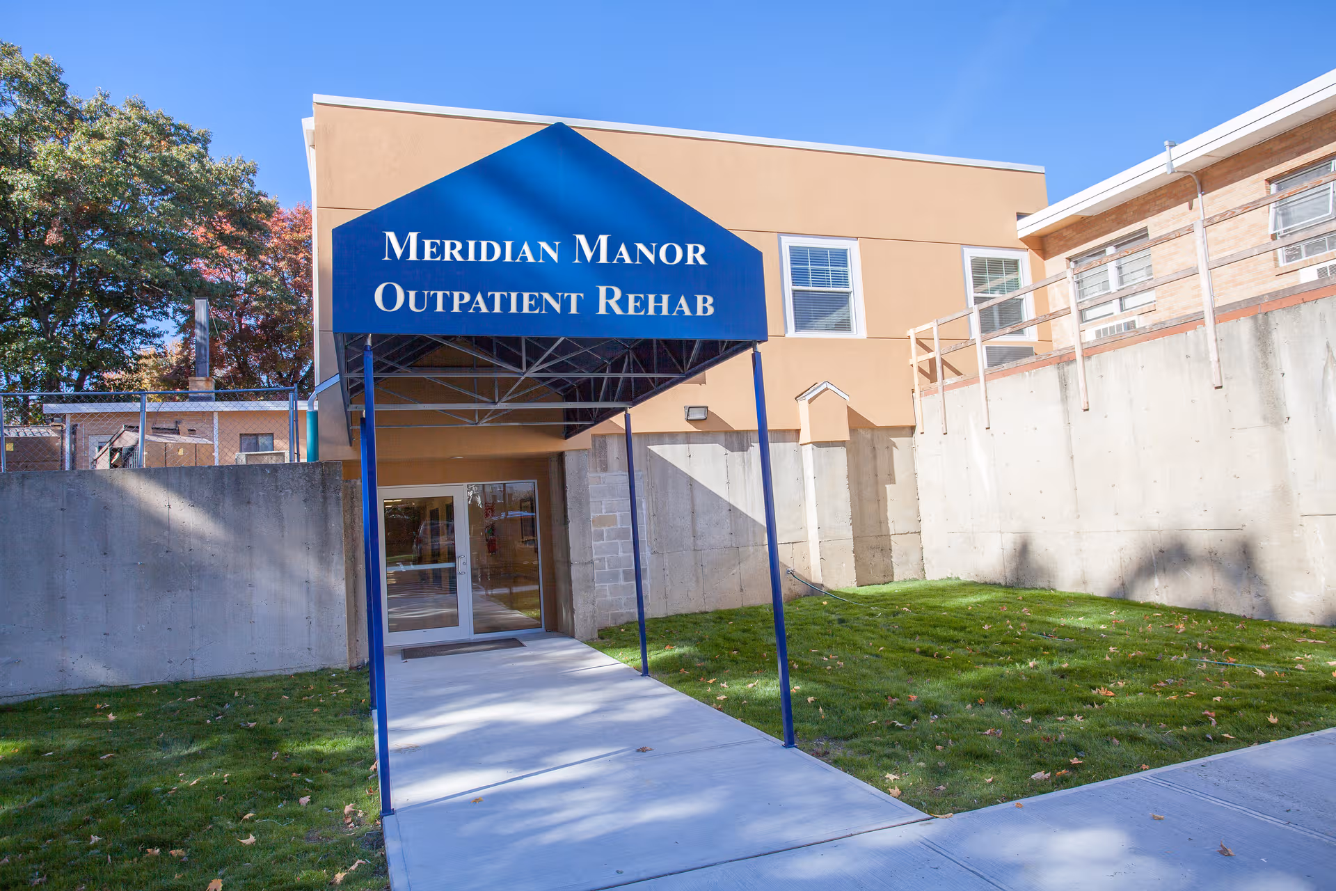 Front entrance of Meridian Manor Outpatient Rehab, with blue canopy, glass doors, and surrounding grassy area.