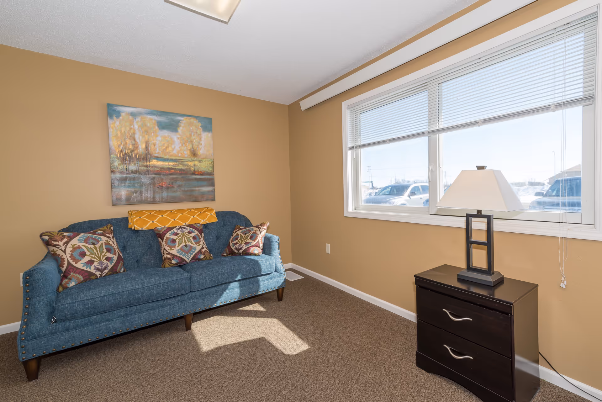 A bright sitting room with a teal sofa adorned with patterned pillows, a landscape painting above, and a lamp on a dark two-drawer table by the window.