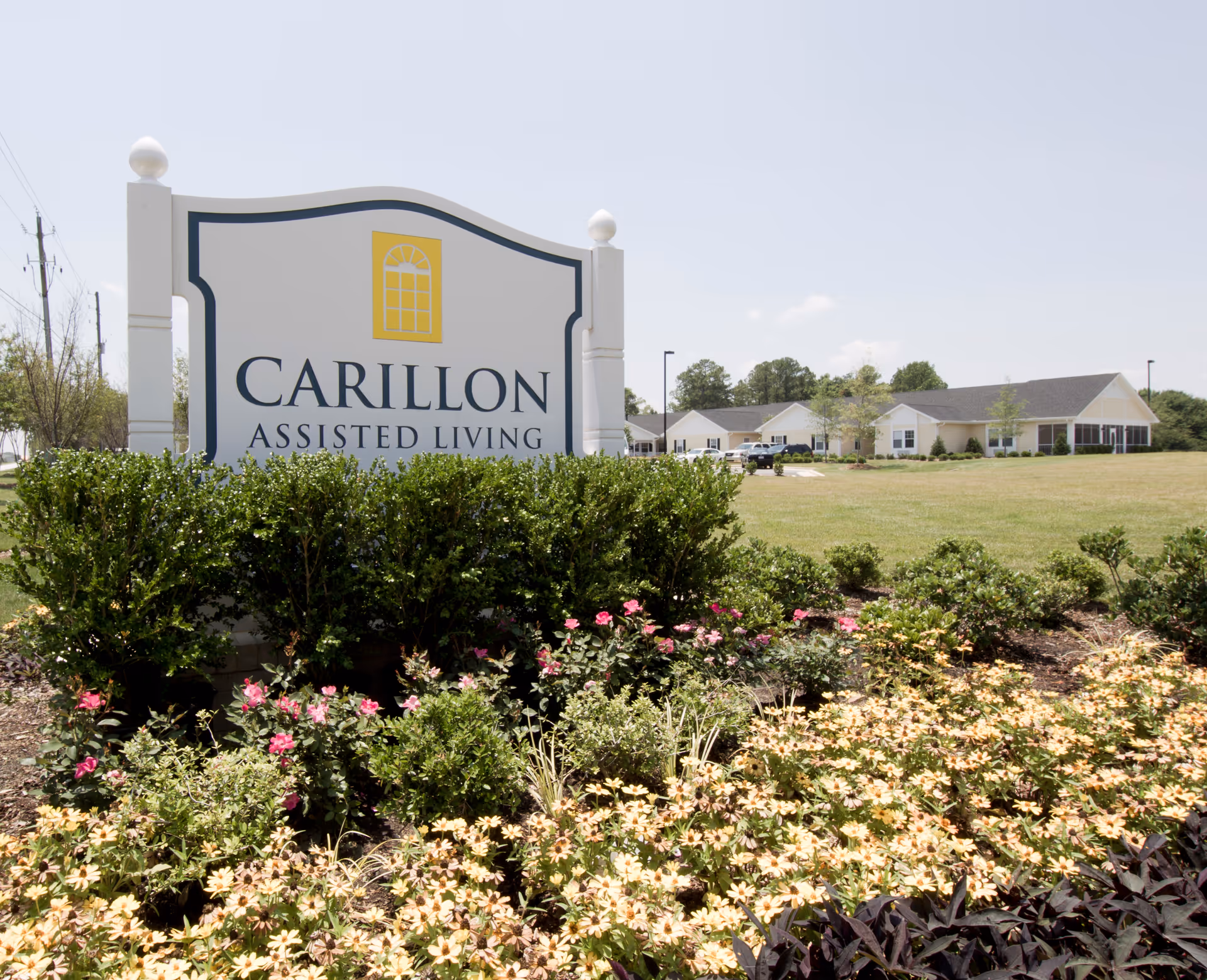 A large white sign with a yellow window logo and the text 'CARILLON ASSISTED LIVING' in front of a landscaped garden with flowers and bushes. In the background, there is a single-story building with a lawn and trees under a clear sky.