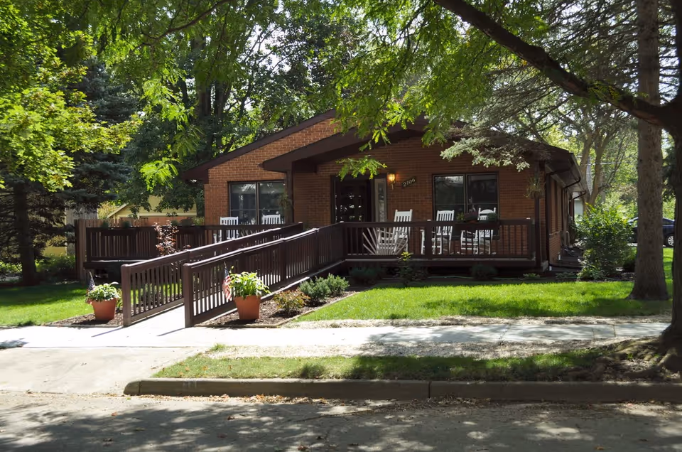 Brick senior living facility with a wooden ramp, porch, and white rocking chairs.