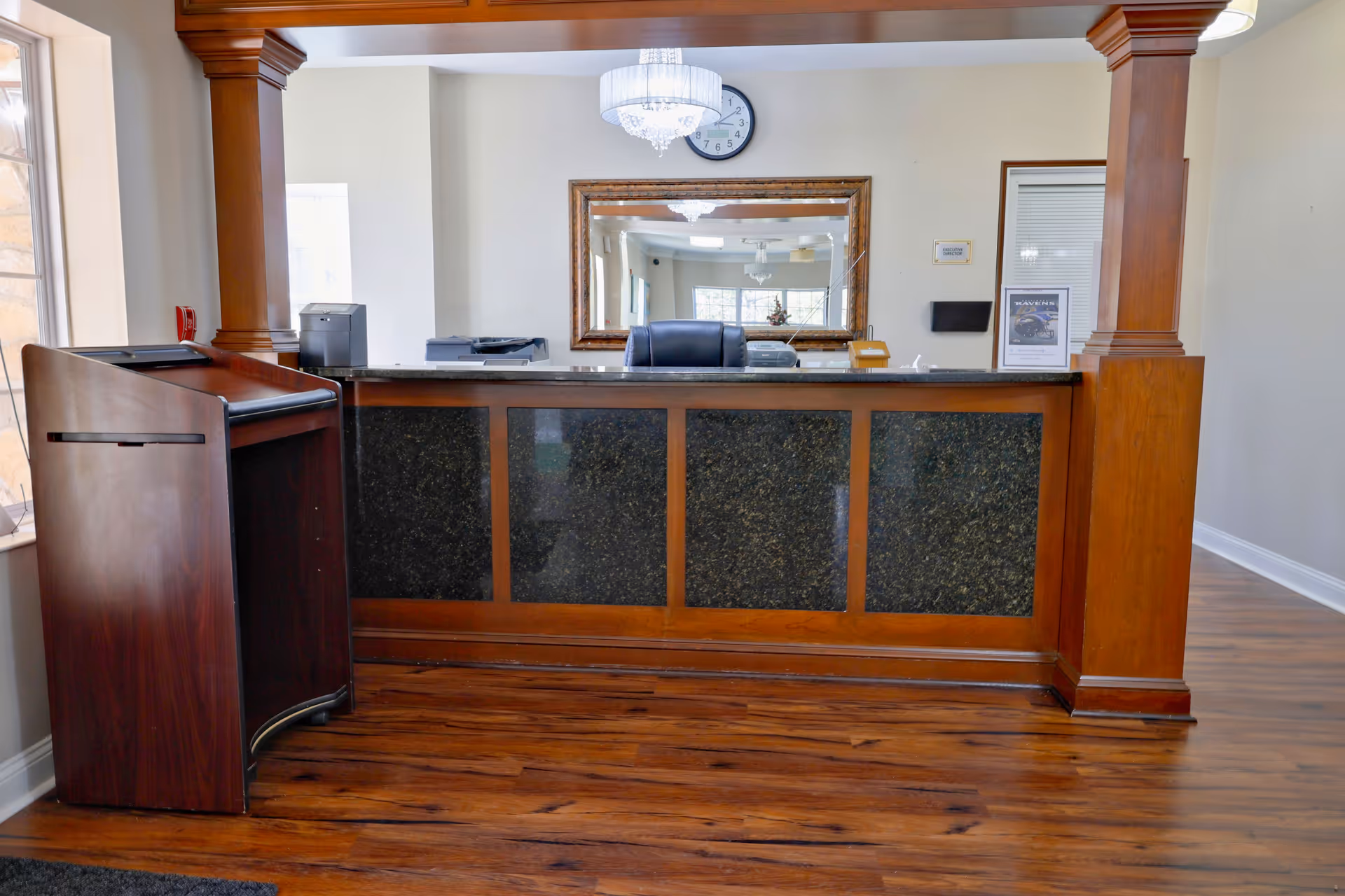 Reception desk area with wooden paneling and black granite front, a black office chair behind the desk, a large mirror on the wall, a clock above the mirror, and a chandelier hanging from the ceiling. The floor is wooden, and there is a wooden podium or stand to the left of the desk.