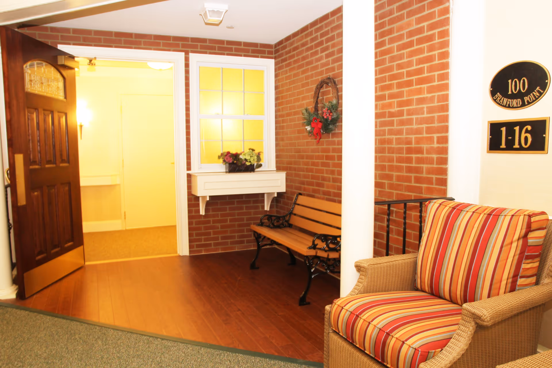 An indoor seating area with a wooden bench and a cushioned armchair with red, orange, and yellow stripes. The walls are brick with a white column and a small white shelf holding a flower pot. A wreath with red decorations hangs on the brick wall. A wooden door is open, leading to a brightly lit hallway. Two black and gold plaques on the wall display the numbers '100 Branford Point' and '1-16'.