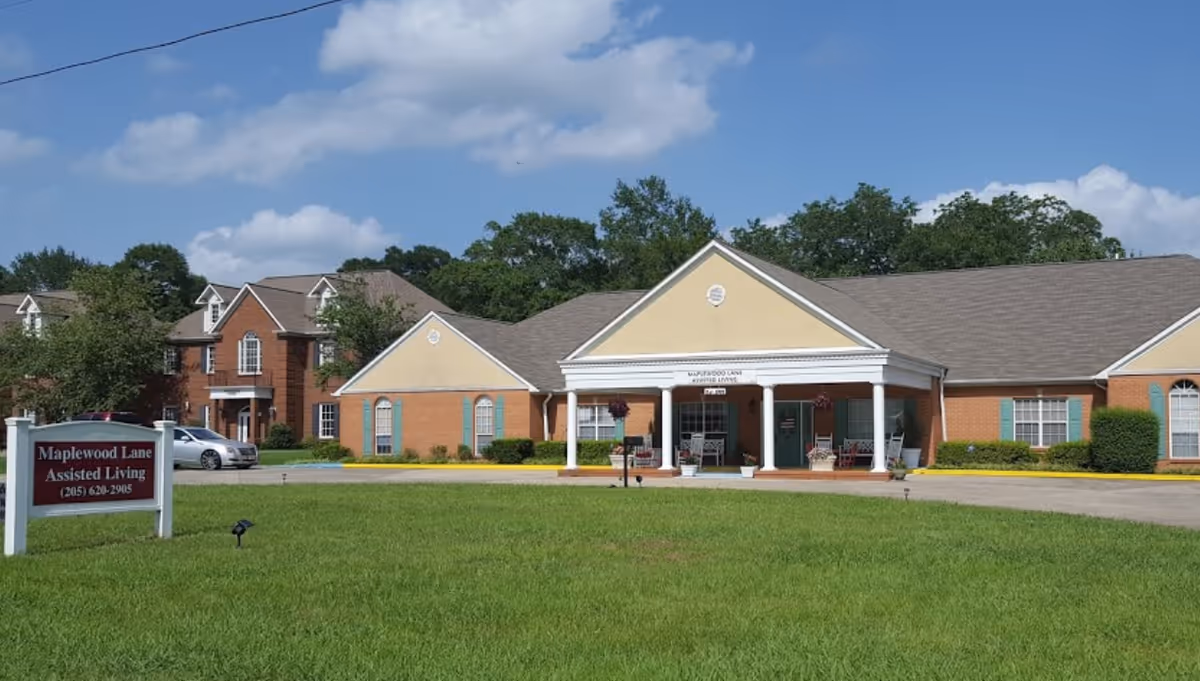 Exterior view of Maplewood Lane Assisted Living facility showing a single-story building with a covered entrance supported by white columns, surrounded by green grass and trees under a blue sky with some clouds. A sign with the facility's name and phone number is visible on the lawn.