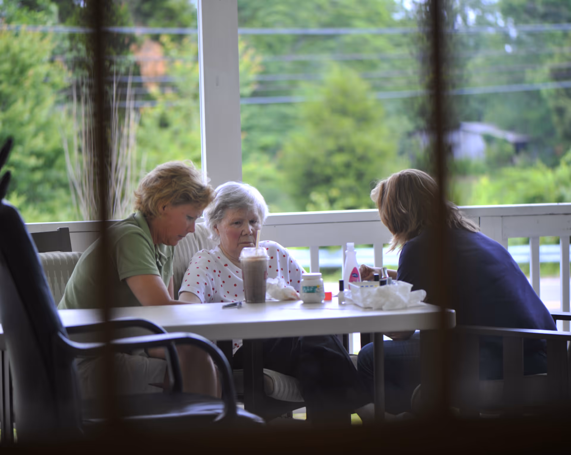 Three women sitting around a table on a porch or patio, engaged in conversation or an activity, with green trees and outdoor scenery visible in the background.