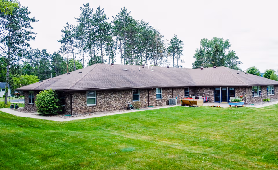 Single-story brick building with a brown roof surrounded by green grass and tall trees in the background under a cloudy sky.