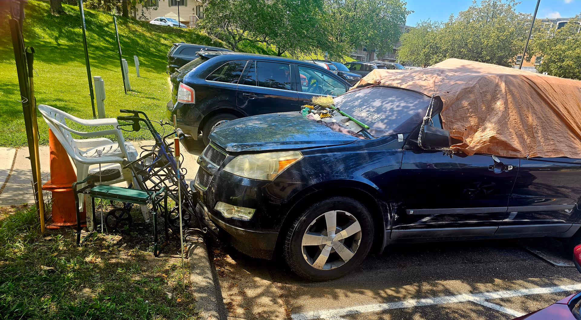 Dark blue SUV with a tarp-covered car nearby and stacked outdoor chairs.