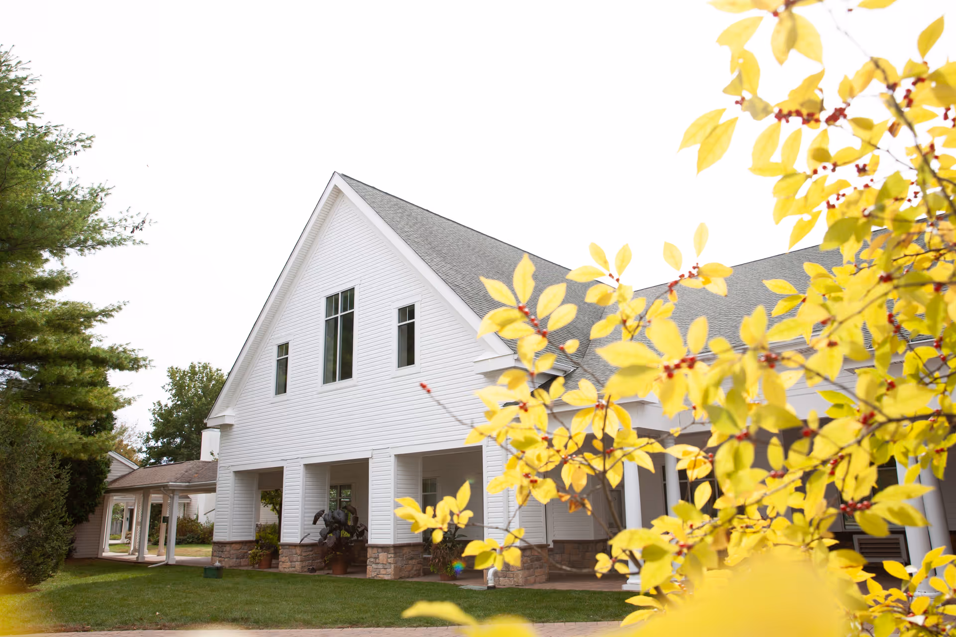 White two-story senior living building with a peaked roof and yellow foliage in foreground.