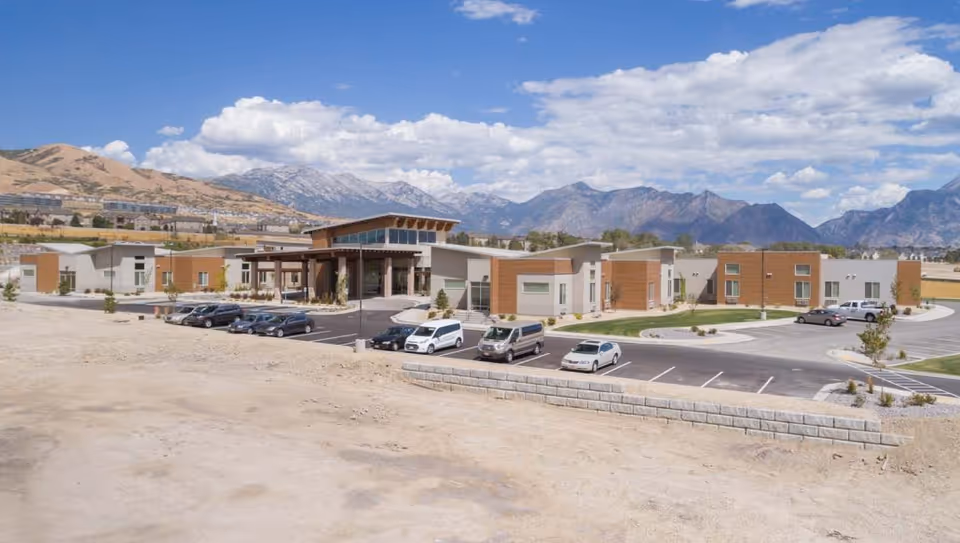 Modern senior living center front with parking lot and mountain backdrop.