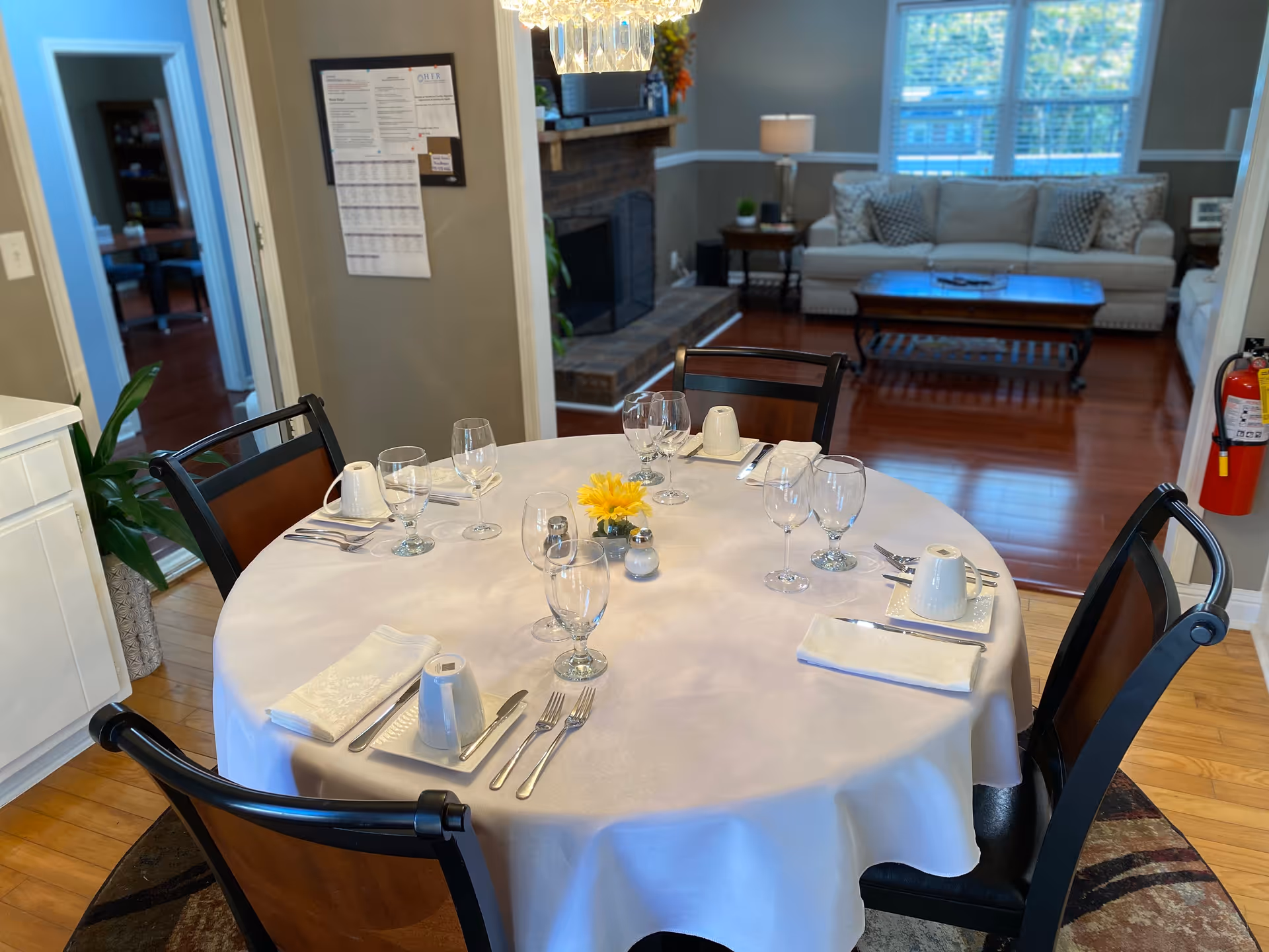 Round dining table with white tablecloth, glassware, and a yellow centerpiece in a bright dining room.