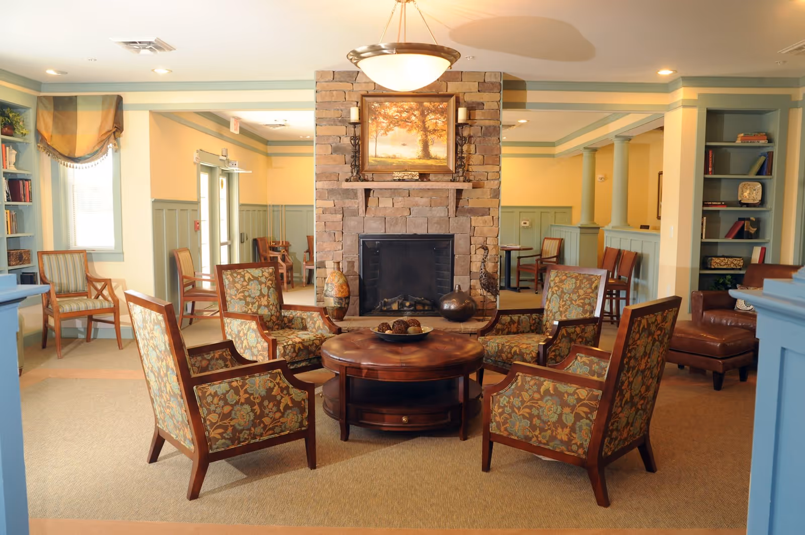 A cozy living room area with a stone fireplace in the center, surrounded by six floral upholstered armchairs arranged around a round wooden coffee table. The room features built-in bookshelves, soft yellow walls with green trim, and warm lighting from ceiling fixtures and a hanging lamp above the fireplace. There are additional chairs and tables in the background near windows and doorways.