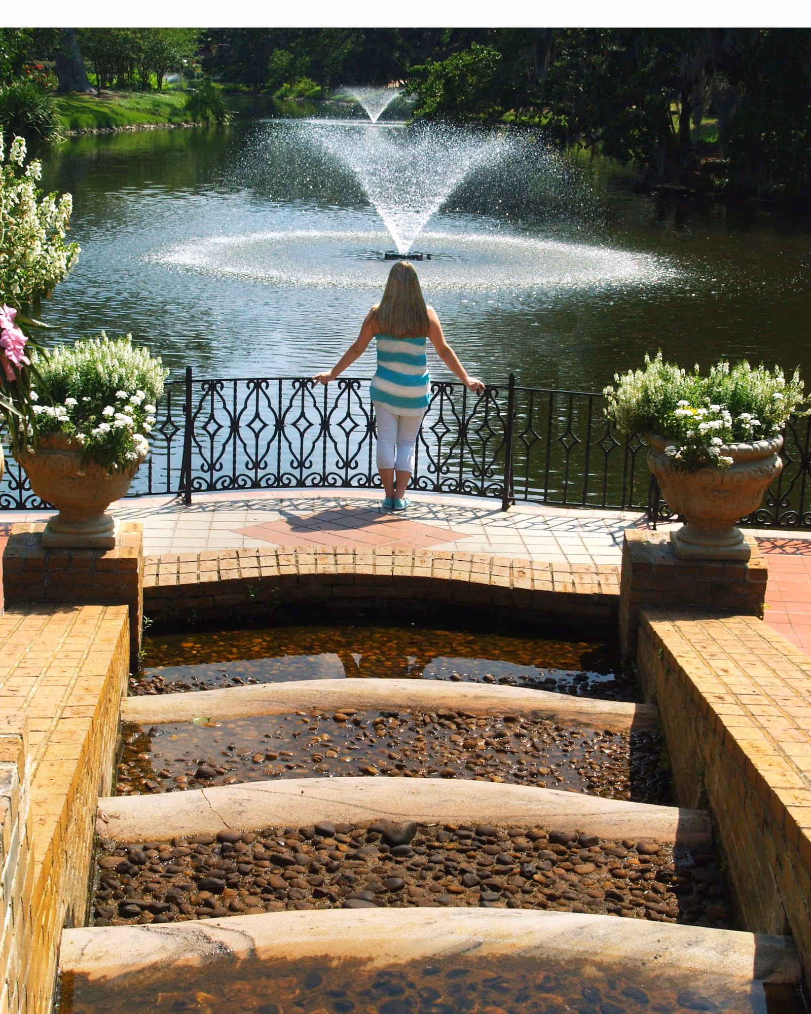 Person stands at a decorative railing overlooking a pond with a fountain in a landscaped garden.