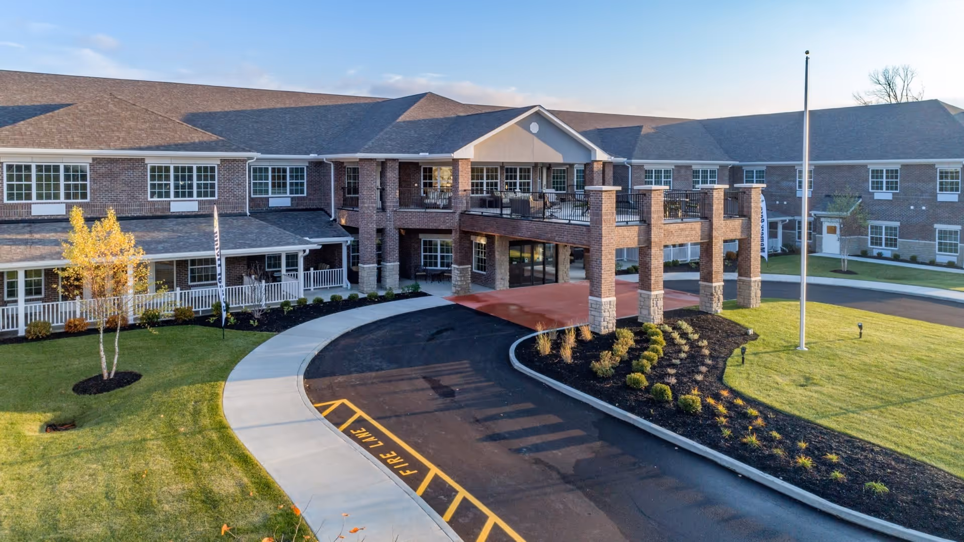 Exterior view of a two-story senior living facility with brick walls and large windows. The building features a covered entrance supported by brick columns, a curved driveway with a fire lane marked, well-maintained green lawns, small trees, and landscaped bushes under a clear sky.