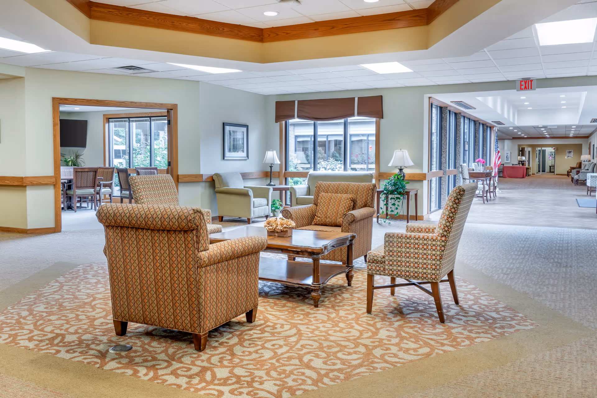 Bright common seating area with upholstered armchairs around a coffee table in a senior living facility hallway.