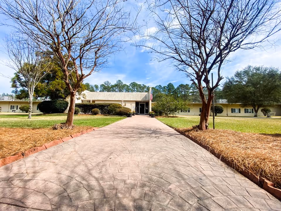 Wide paved walkway leading to the entrance of a single-story building with an American flag near the door, surrounded by grass, trees, and shrubs under a partly cloudy blue sky.