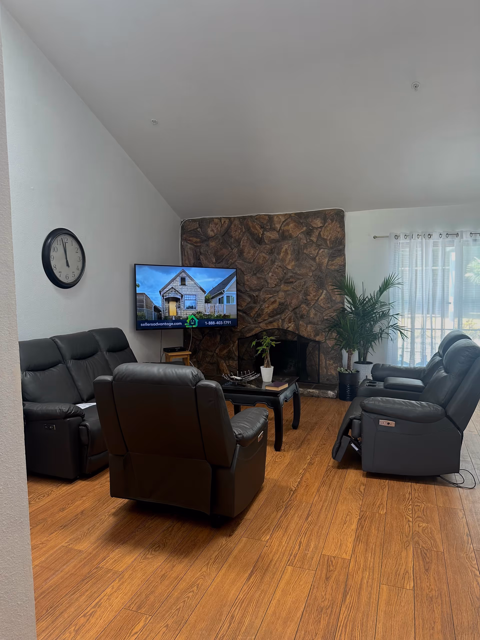 A living room with three black leather recliners arranged around a black coffee table. The room features a stone fireplace, a wall-mounted flat screen TV displaying a house advertisement, a wall clock showing 11:55, wooden flooring, and a large window with sheer white curtains letting in natural light. There are also two potted plants near the fireplace.