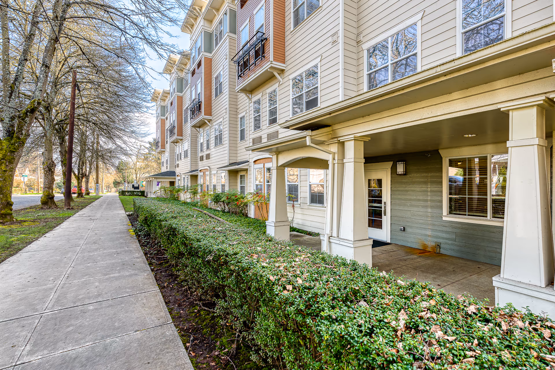 Covered entrance and facade of a multi-story senior living building with a sidewalk, hedges, and trees.