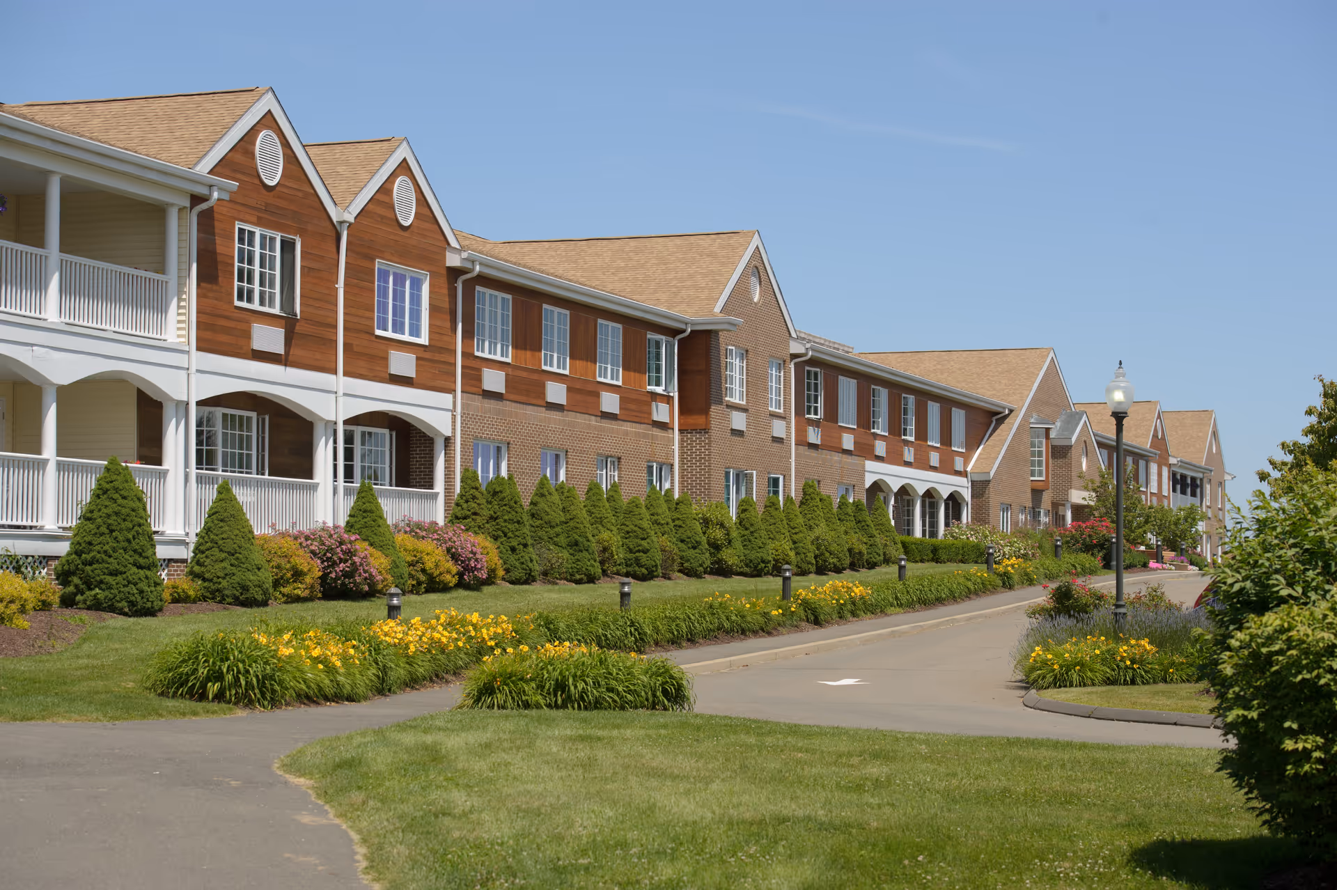 Exterior view of a long two-story senior living building with porches, landscaped shrubs and a driveway.