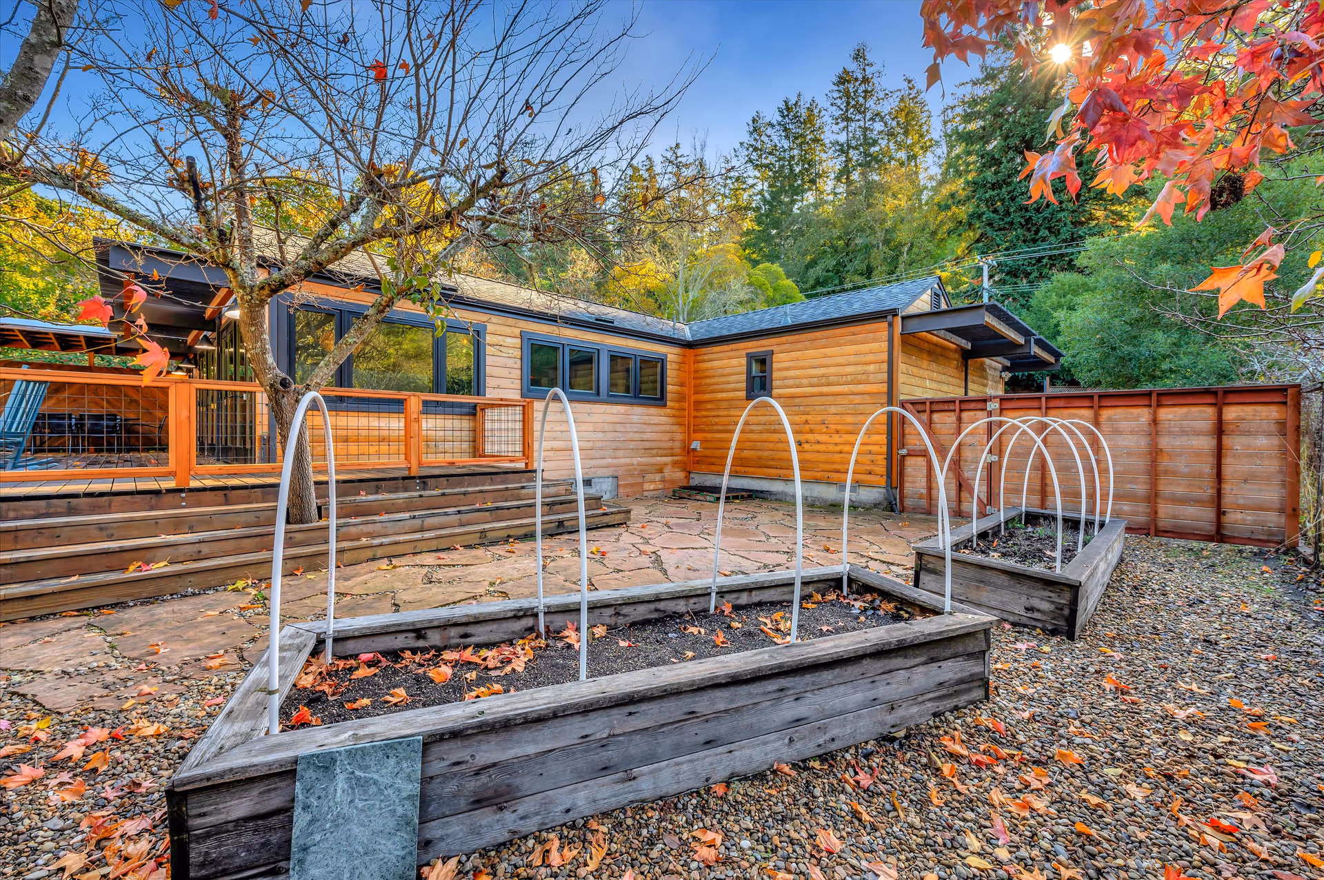 Wood-paneled single-story building with a fenced deck and multiple raised garden beds in a leafy outdoor courtyard during autumn.