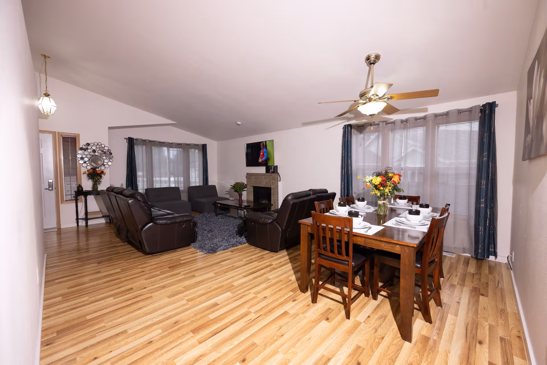 A spacious living and dining area with wooden flooring. The living room features dark leather sofas arranged around a coffee table on a gray rug, with a wall-mounted TV above a fireplace. The dining area has a wooden table set for six with white dishes and a vase of colorful flowers. Large windows with sheer curtains and blue drapes allow natural light to fill the room. A ceiling fan with lights is mounted above the dining table.