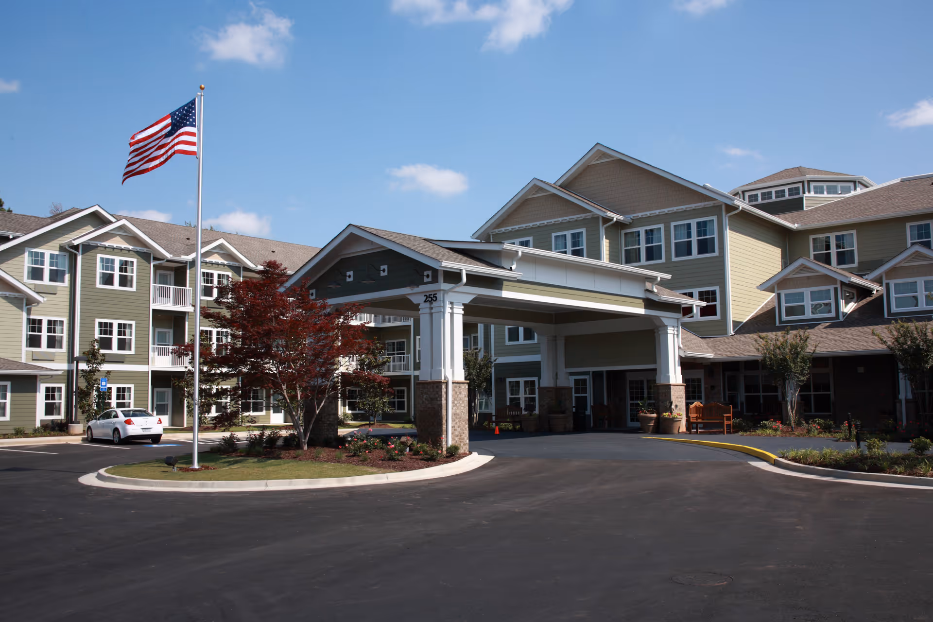 Front exterior view of a multi-story senior living facility with a covered entrance, an American flag on a flagpole, landscaped greenery, and a clear blue sky.