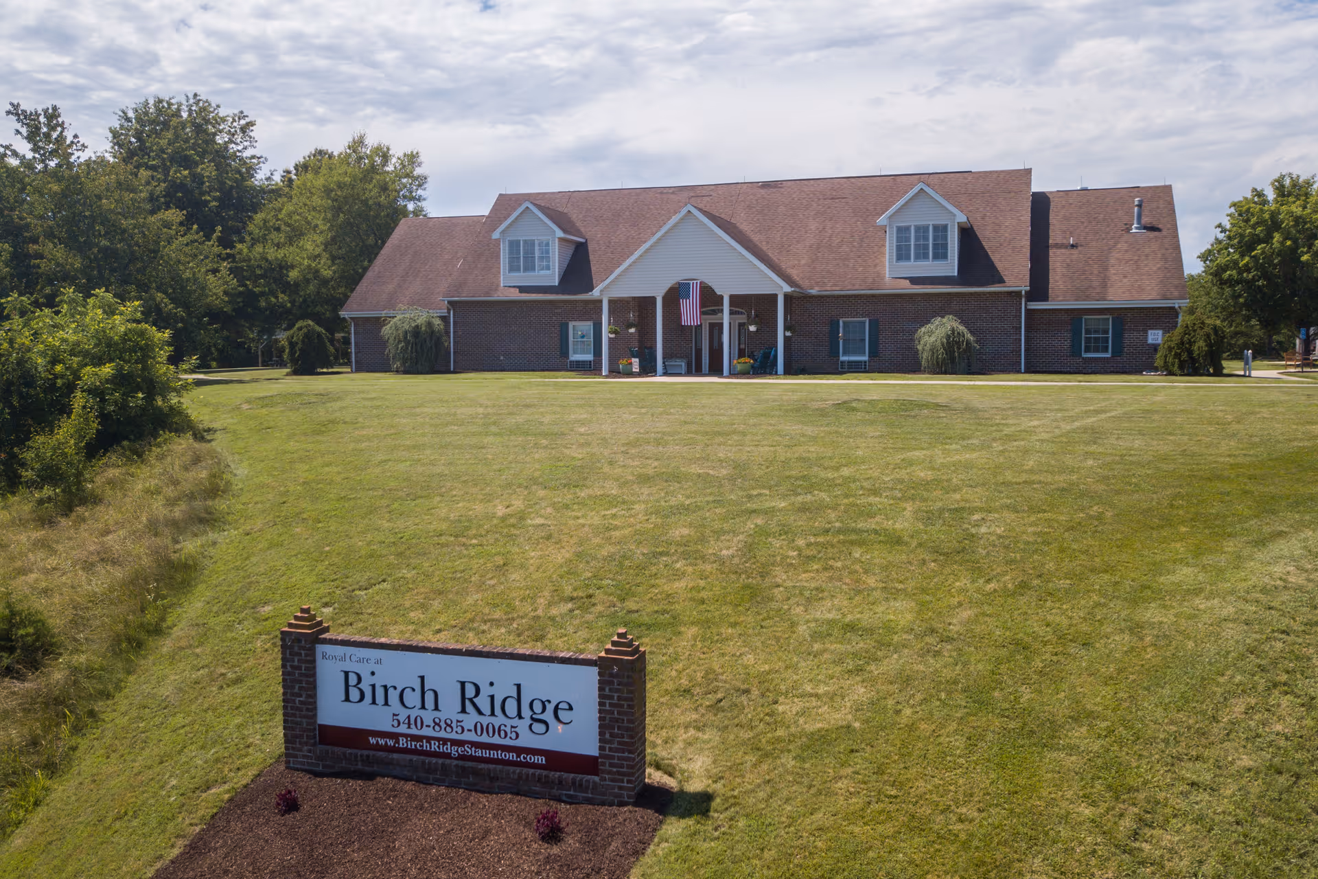 A single-story brick building with a brown roof and white trim, featuring two dormer windows and a covered entrance with an American flag. The building is set on a large grassy lawn with trees in the background. In the foreground, there is a brick sign that reads 'Royal Care at Birch Ridge 540-885-0065 www.BirchRidgeStaunton.com'.