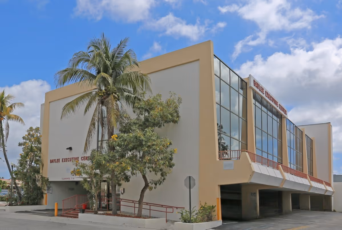 Two-story beige office building with large glass windows, palm trees, and a blue sky.