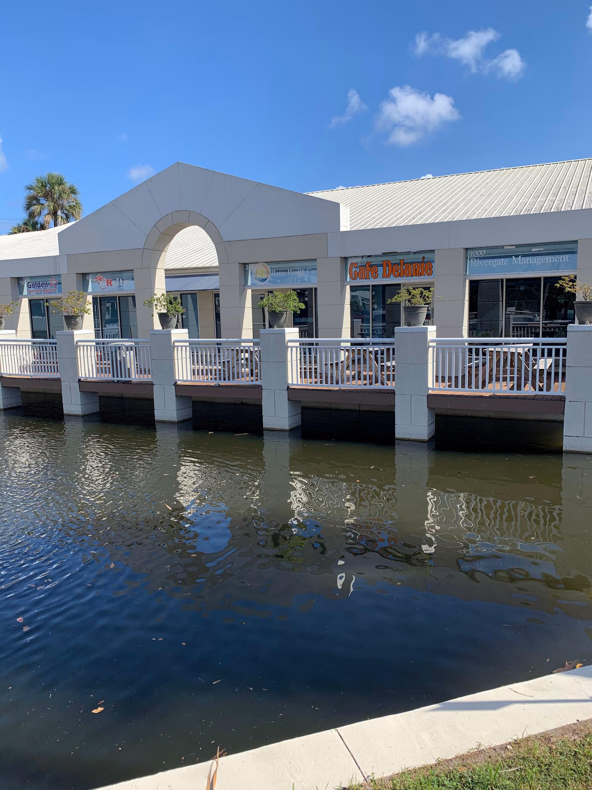 White building with arched entrance along a canal, storefronts, railing, under blue sky.