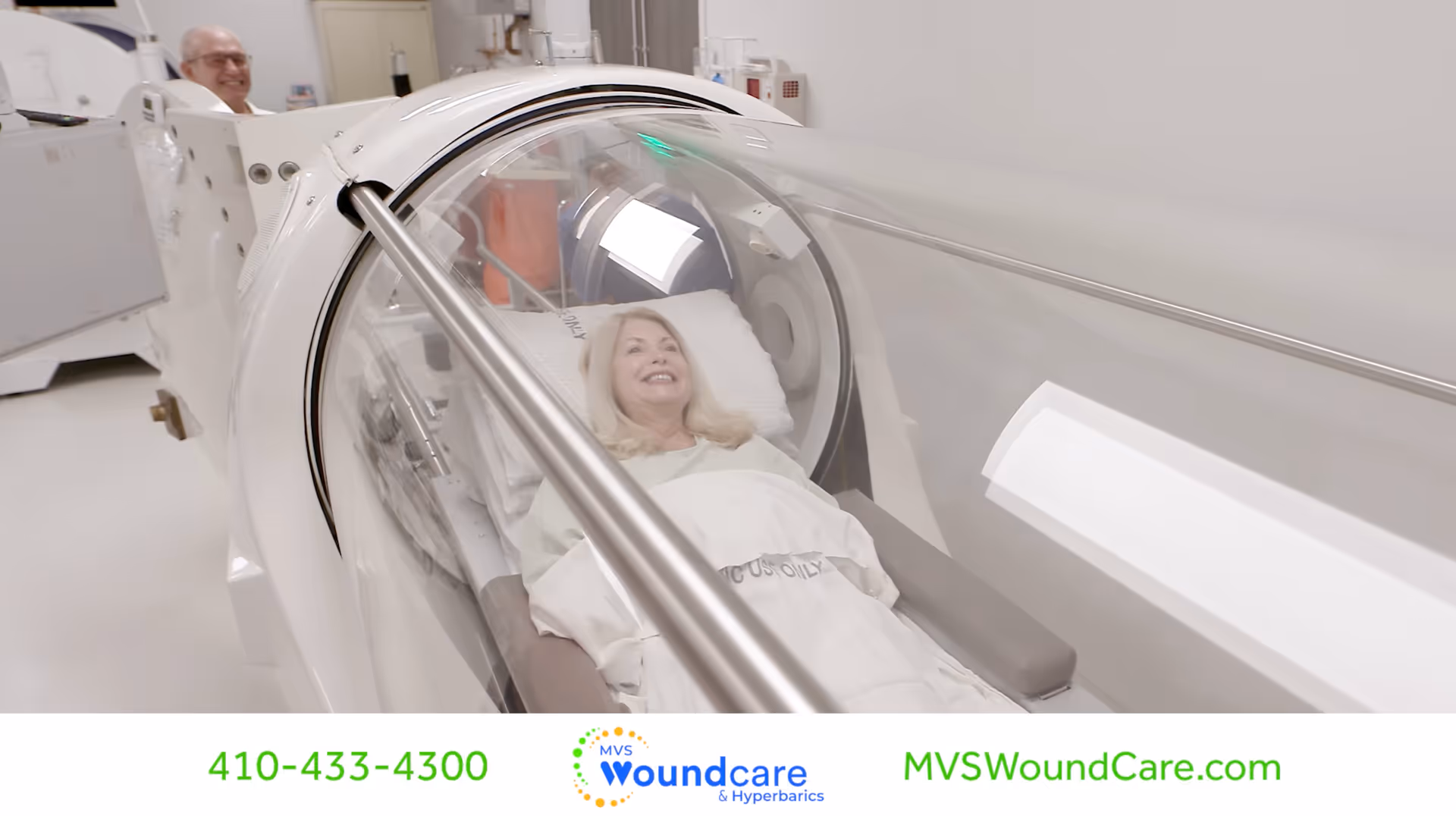 Patient lies in a hyperbaric chamber while a smiling staff member stands nearby.