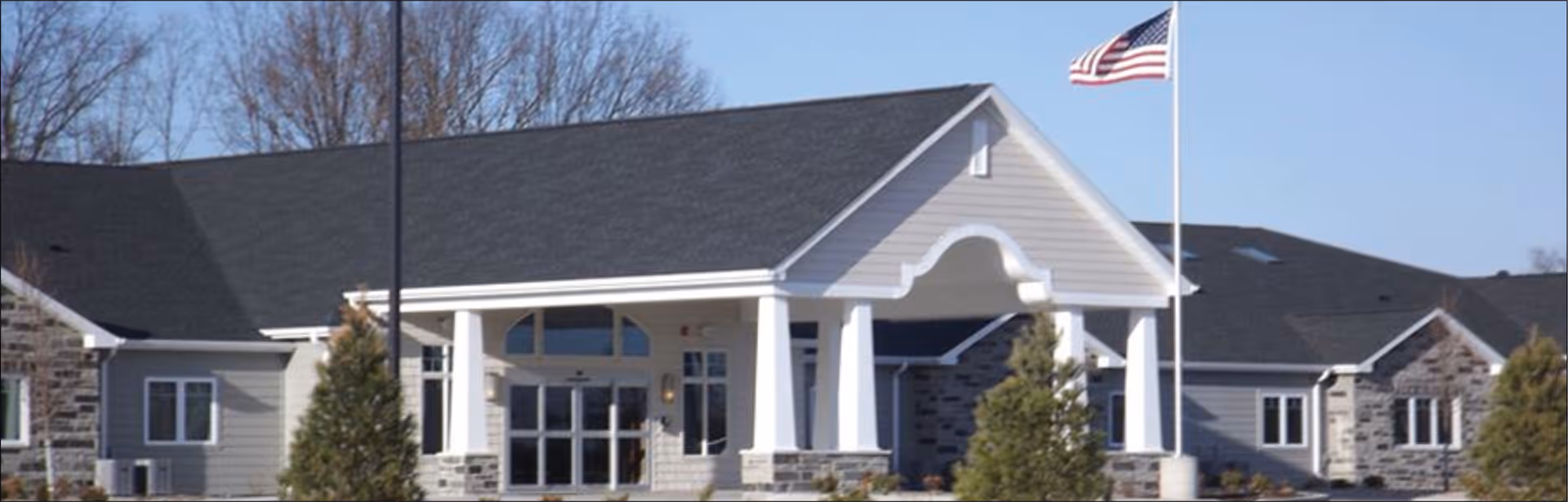Exterior view of a senior living facility building with a covered entrance supported by white columns, stone and siding facade, and an American flag flying on a flagpole in front.