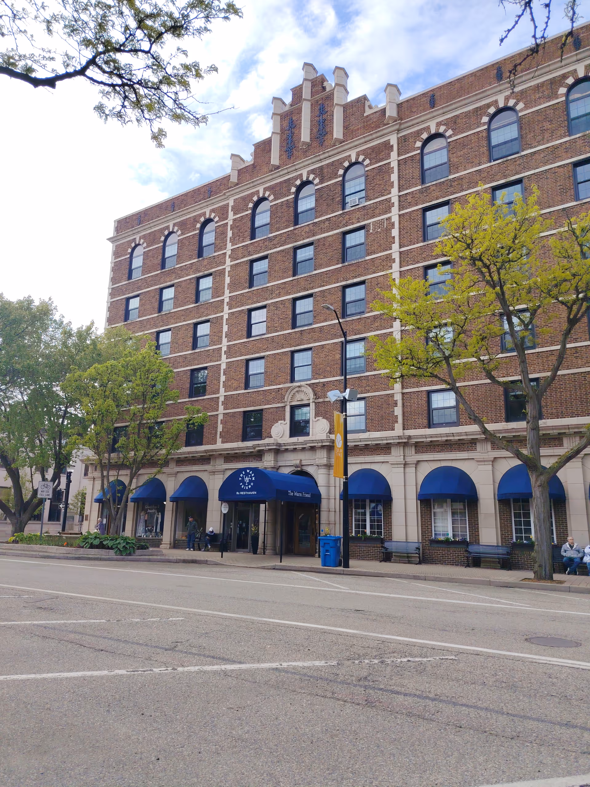 Brick senior living facility front with blue awnings, trees, and entrance.