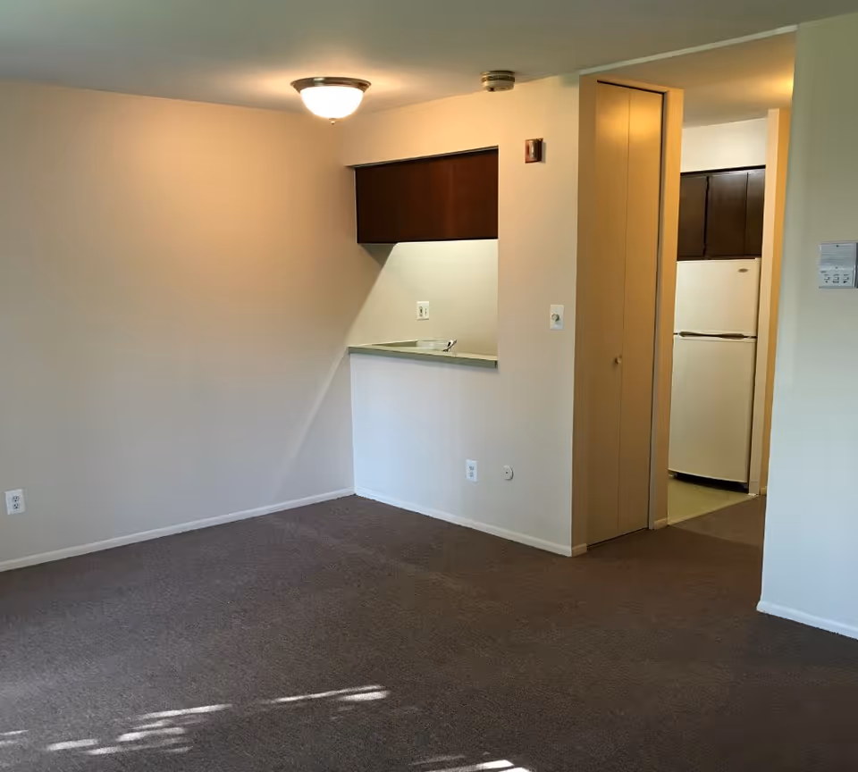 Empty carpeted living room with a pass-through countertop to a small kitchen and a refrigerator visible through the doorway.