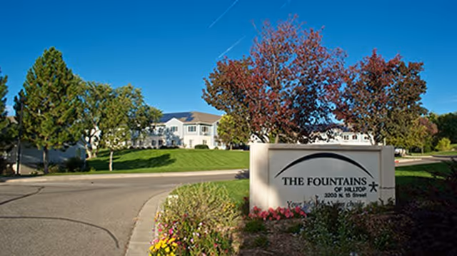 Exterior view of The Fountains of Hilltop with sign, road, and trees.
