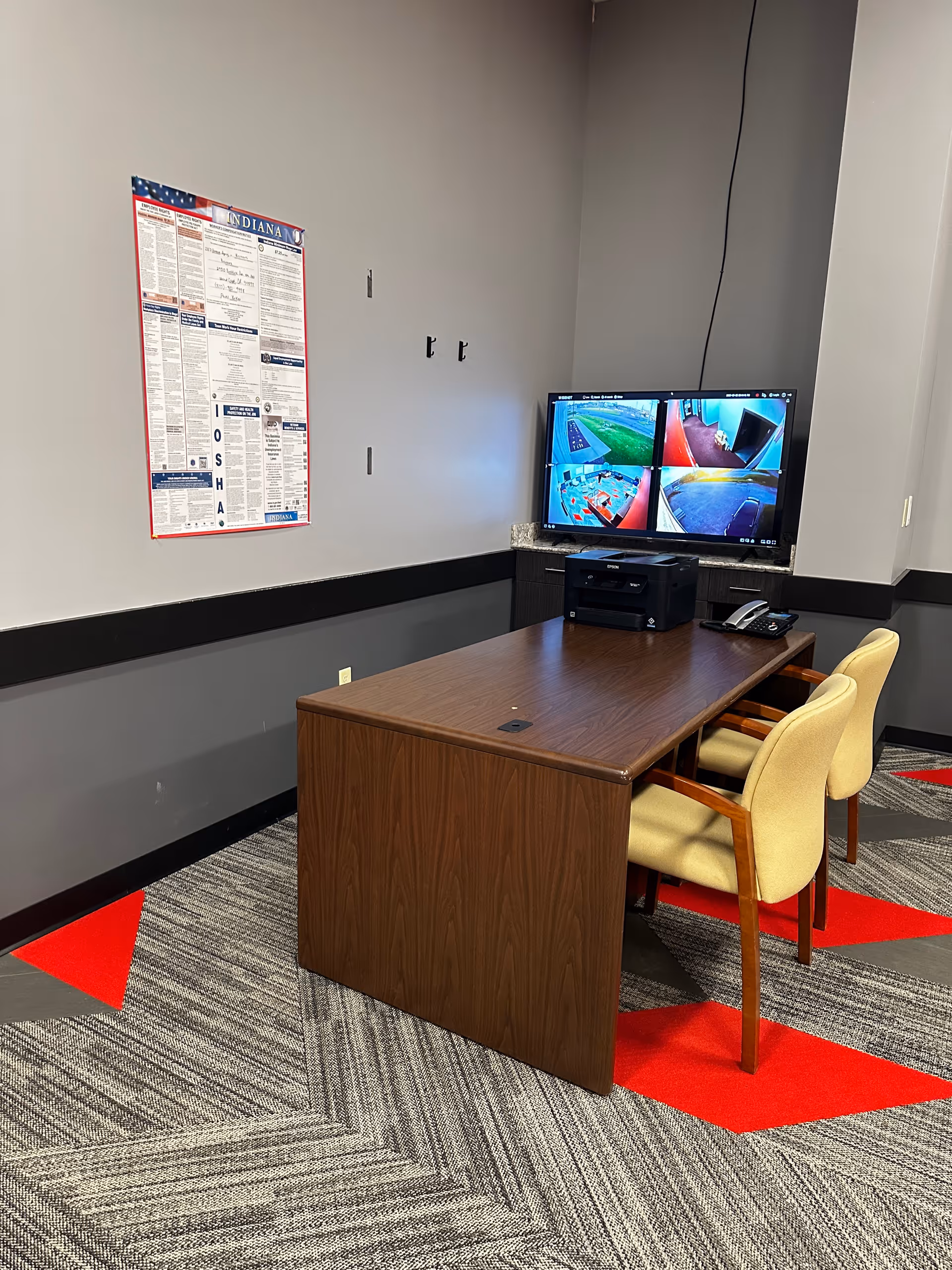 Interior office area with a wooden desk, beige chairs, and a six-camera security monitor.