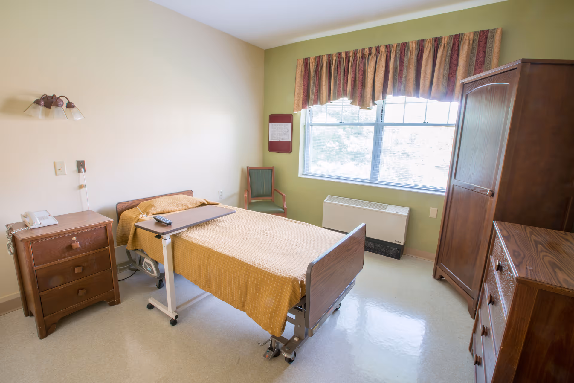 A simple bedroom in a senior living facility with a single hospital-style bed covered with a mustard yellow blanket, a wooden nightstand with a telephone, a green chair near a window with patterned curtains, a wooden wardrobe, and a wooden dresser. The room has light-colored walls and a linoleum floor.