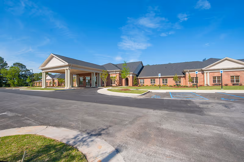 Exterior view of a single-story brick building with a covered entrance supported by columns, surrounded by a parking lot with marked handicap spaces and some landscaping under a clear blue sky.