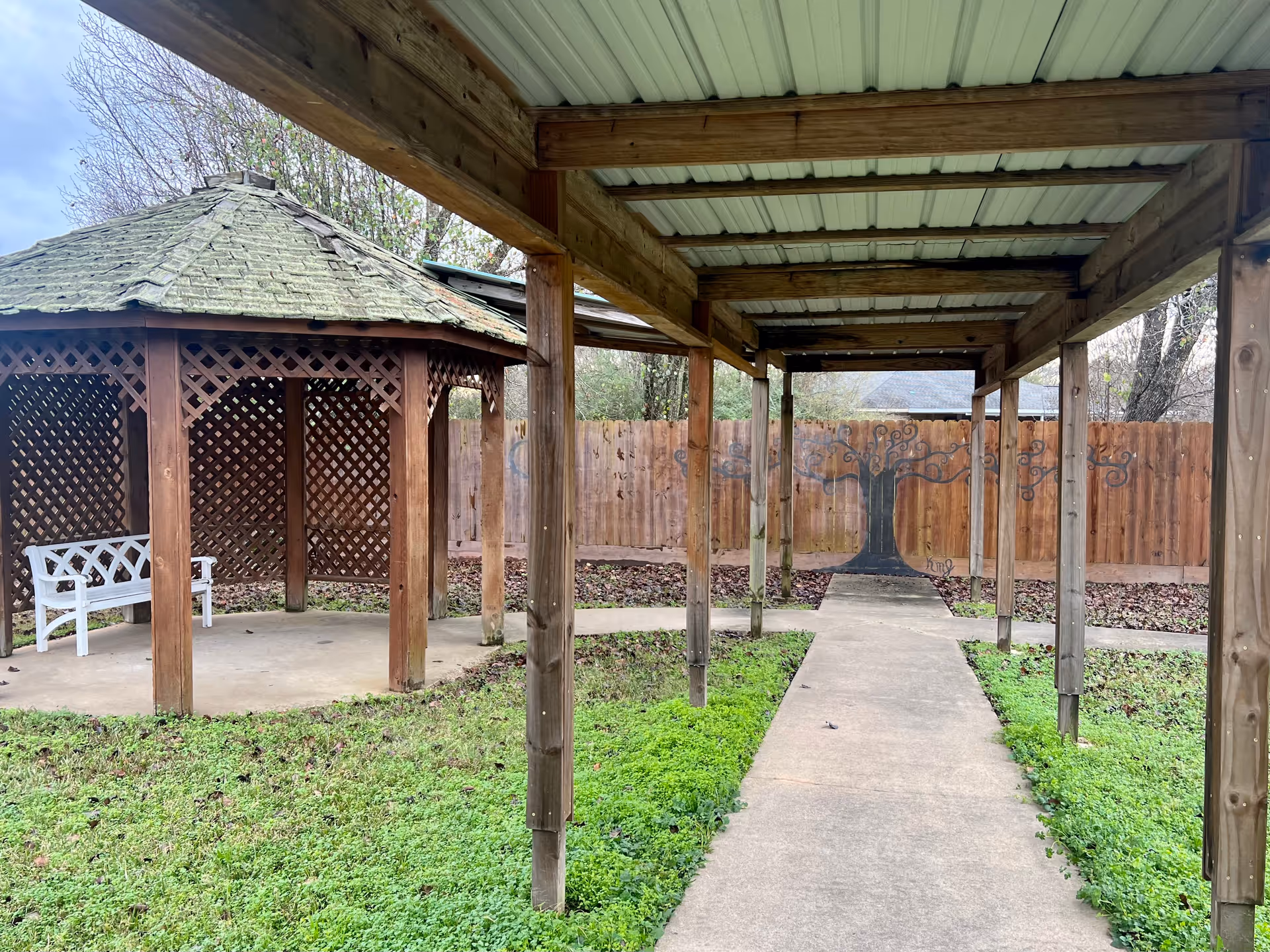 Outdoor area with a wooden gazebo featuring lattice walls and a white bench inside. A covered walkway with wooden posts and a metal roof leads to a wooden fence with a painted tree mural. The ground is covered with green grass and some fallen leaves.