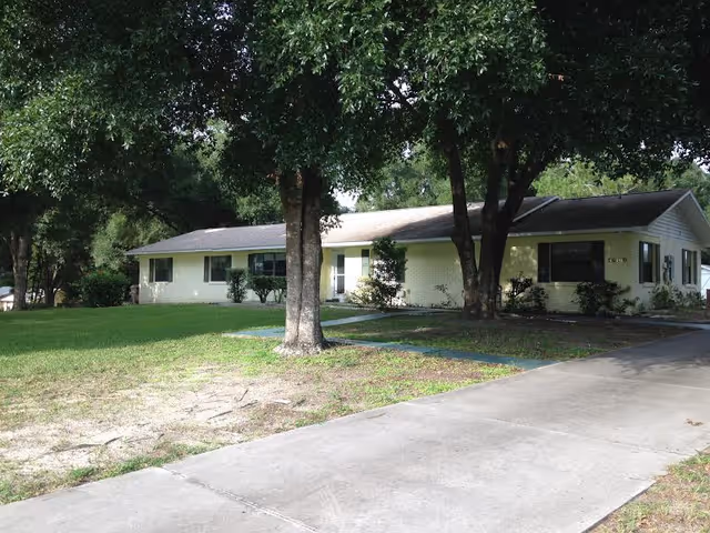 Single-story light-colored house front with a driveway, lawn, and large trees.