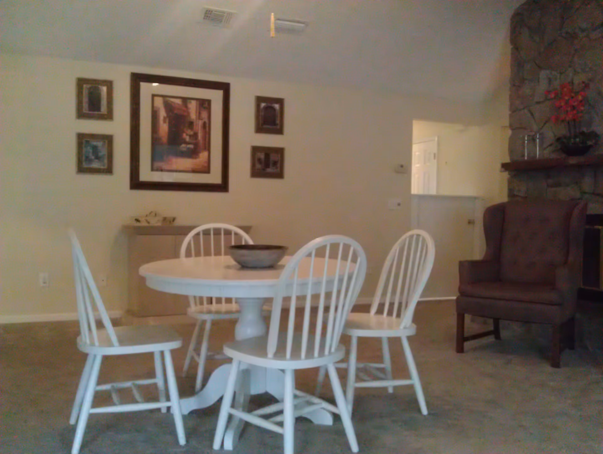 A dining area with a round white table surrounded by four white wooden chairs. On the table is a decorative bowl. Behind the table, there is a wall with five framed pictures arranged around a larger central picture. To the right, there is a brown upholstered armchair next to a stone fireplace with a small plant on the mantel. The room has beige carpet and light-colored walls.