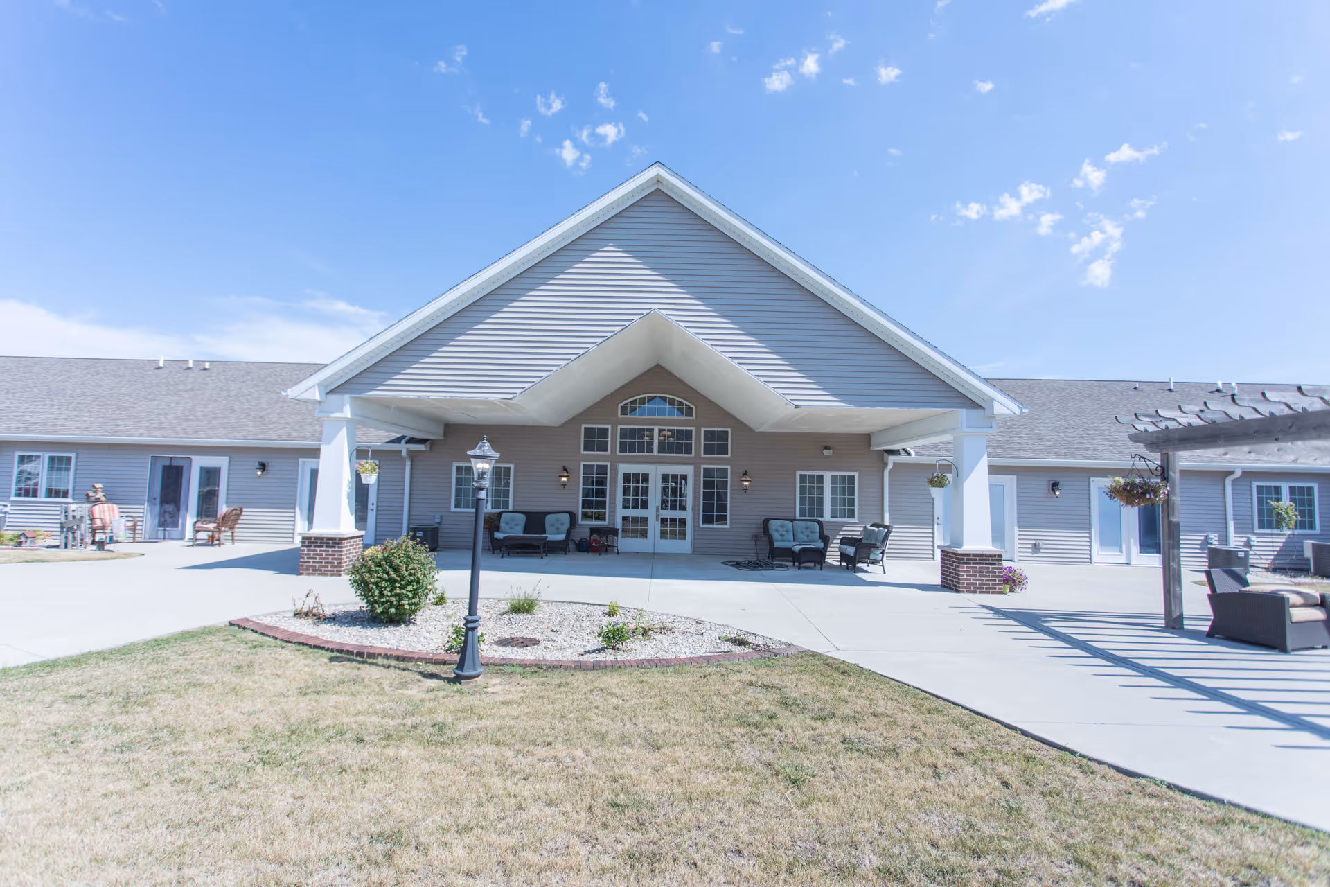 Exterior view of the Villas of Holly Brook Shelbyville building entrance with a covered porch area featuring outdoor seating, a lamp post, and a small landscaped garden in front. The sky is clear with a few clouds.
