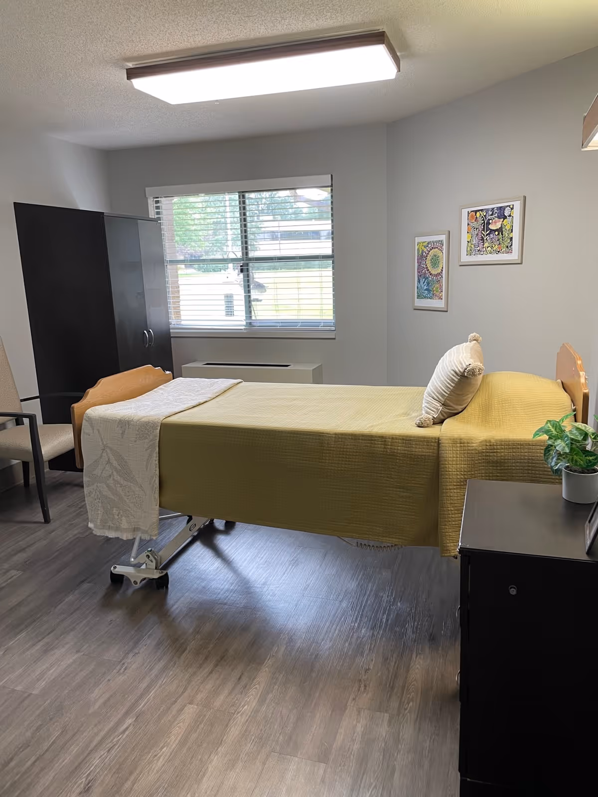 A simple bedroom with a single bed covered in a yellow bedspread and a white pillow. There is a black nightstand with a small green plant on it next to the bed. A window with blinds lets in natural light, and two colorful framed artworks hang on the wall. A black wardrobe and a beige chair are also visible in the room with wood-style flooring.