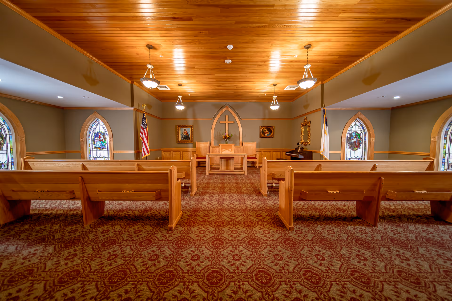 Interior view of a chapel or worship room with wooden pews, a wooden ceiling, stained glass windows, an American flag, a Christian flag, and a cross mounted on the wall behind a small altar with chairs.