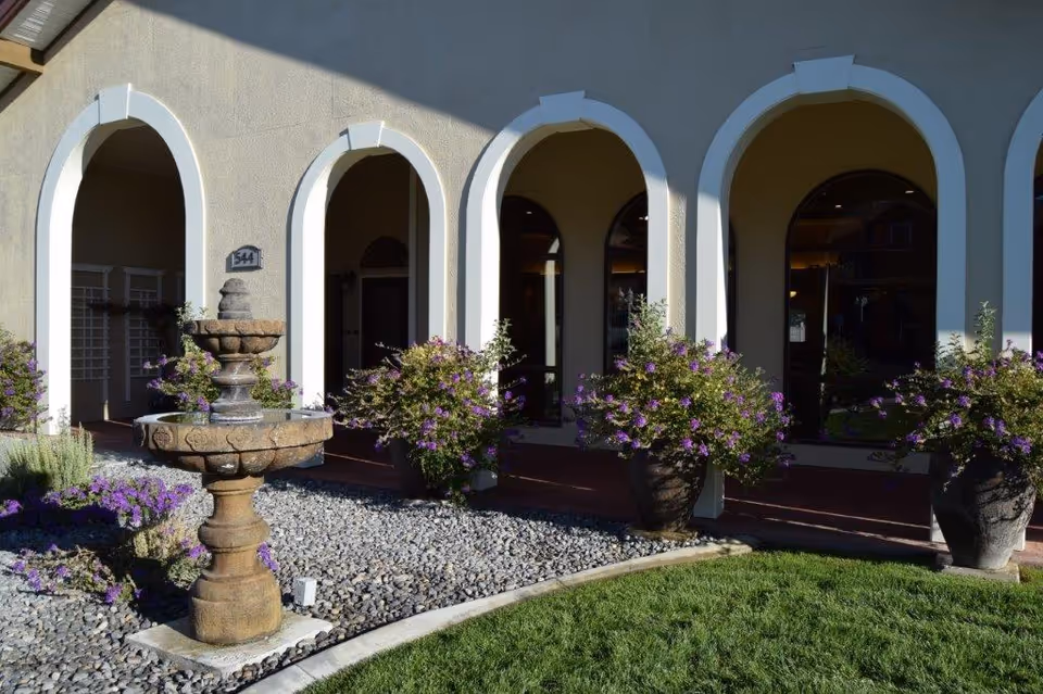 Front of arched building entrance with fountain and purple potted flowers.