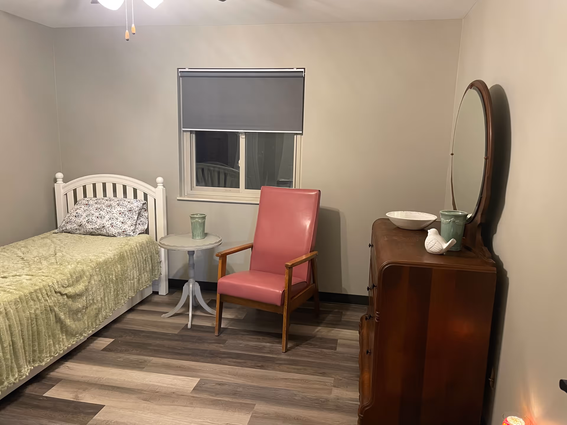 A small bedroom with a white bed, pink chair, dresser with mirror, and window shade.