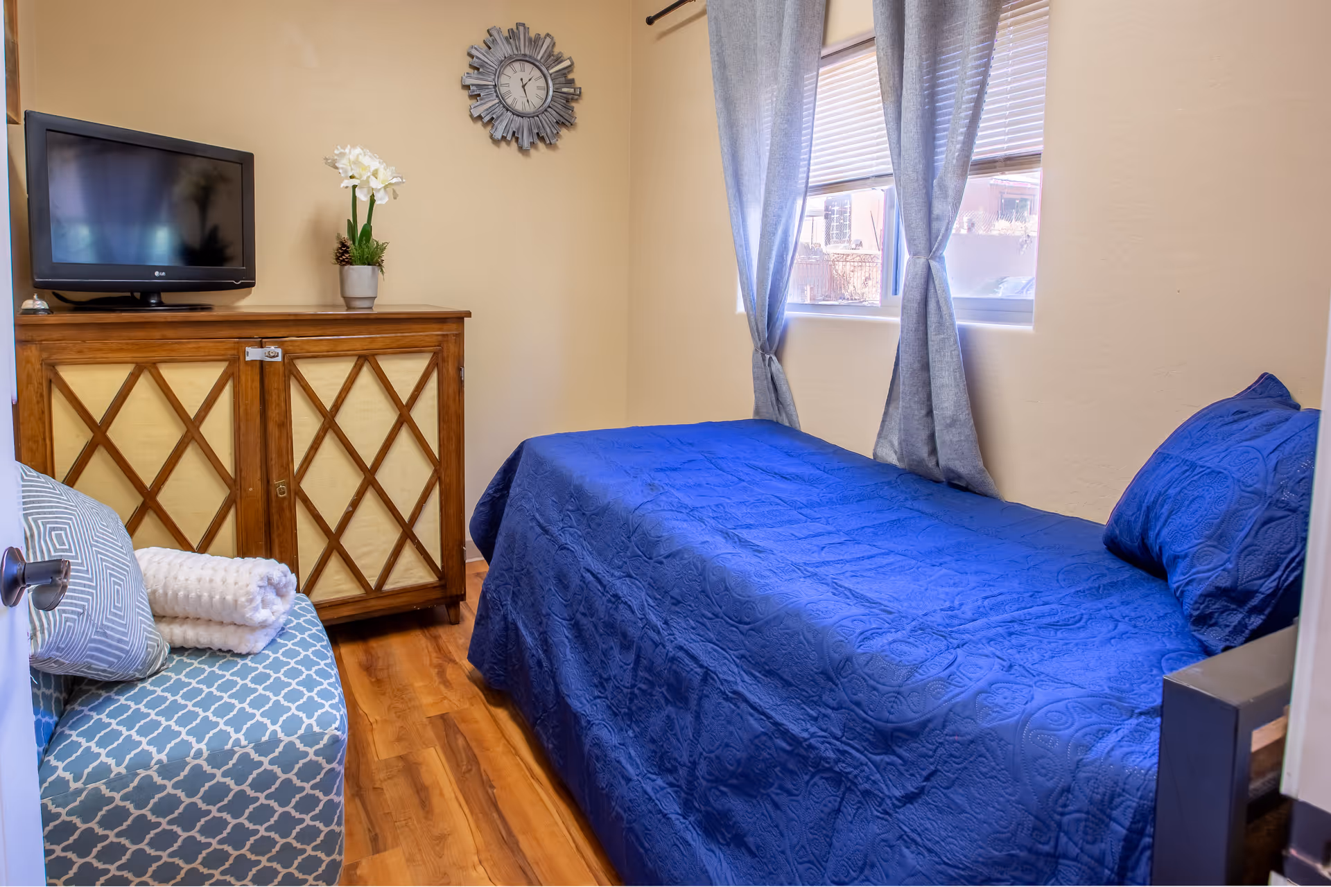 Cozy bedroom in a senior living facility with a blue bed, dresser, TV, and window curtains.