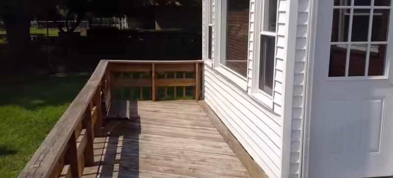 Wooden outdoor deck adjacent to a white-siding building, with a door and green lawn nearby.