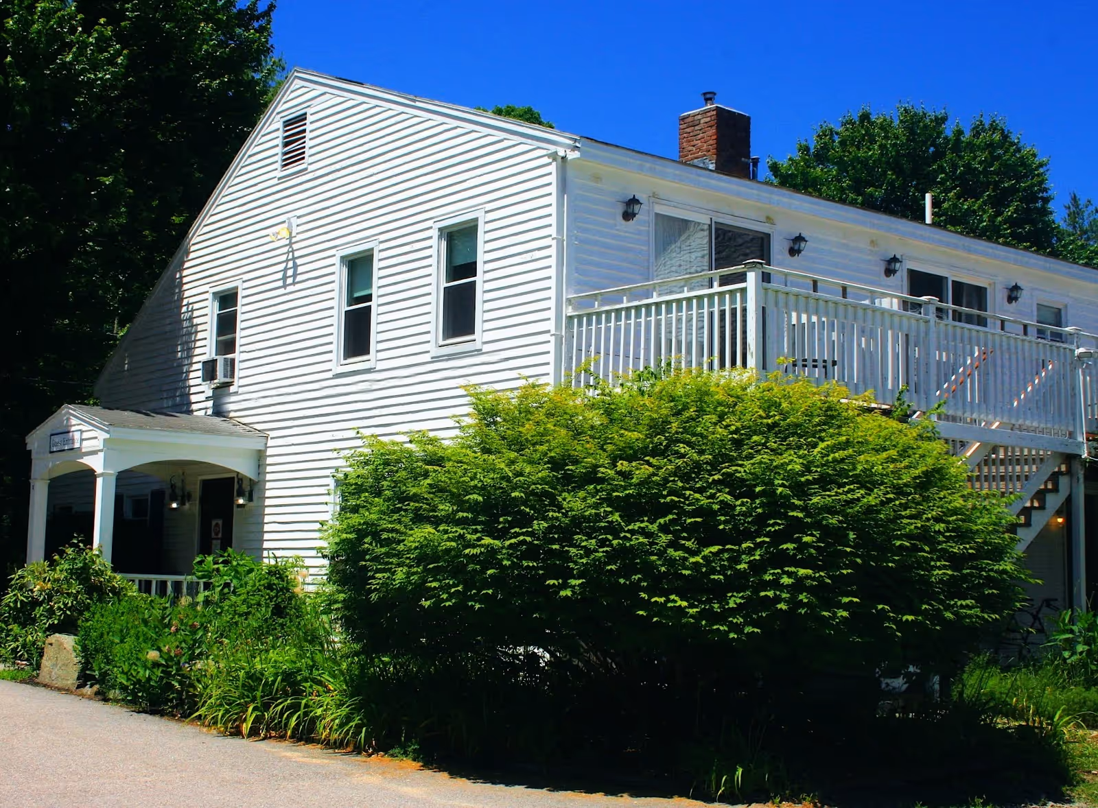 Exterior view of a white two-story building with a porch and balcony, surrounded by green bushes and trees under a clear blue sky.